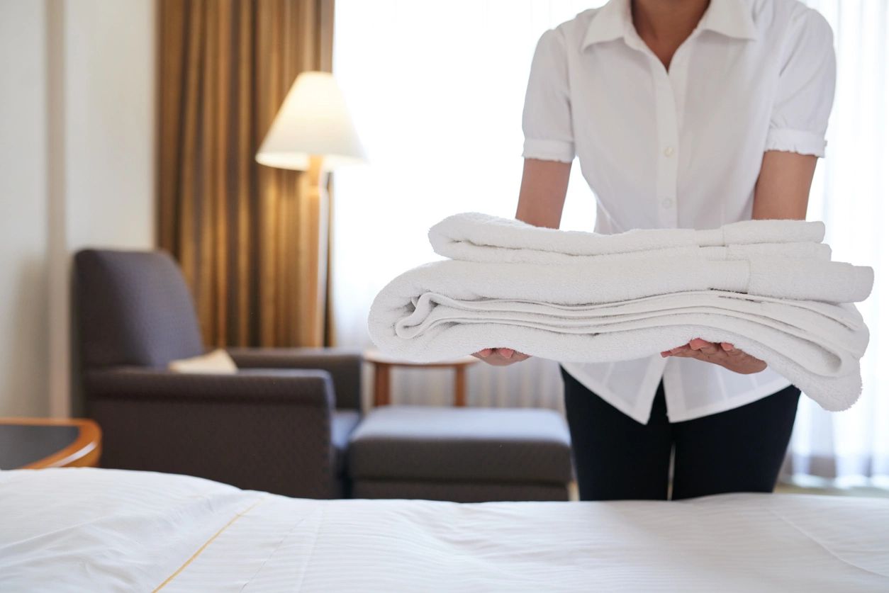 A person holding folded sheets in front of a bed.